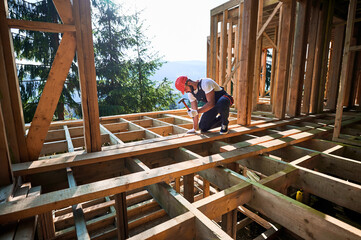 Carpenter constructing wooden frame, two-story house near forest. Bearded man hammering nails with...