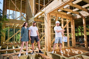 Man designer with plan in hand shows to couple the process of constructing wooden framed house....