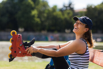 A joyful woman prepares to skate by fastening her roller skates while seated on a bench in a sunny park, expressing happiness and active lifestyle.