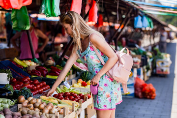 A young woman in a floral dress explores a vibrant outdoor market, selecting fresh vegetables like...
