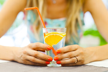 Close-up of a woman's hands holding a glass of water garnished with a fresh orange slice,...