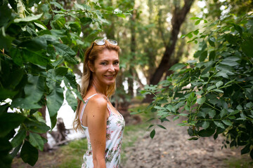 A happy middle-aged woman in a floral dress smiles while walking through a beautiful, green forest, surrounded by fresh foliage.