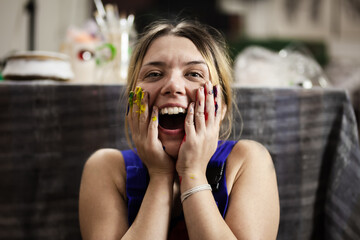 Delighted young female artist with colorful paint smudges on her hands and face, showcasing her...