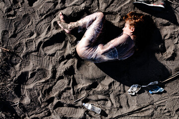 Woman wrapped in plastic lying on a muddy beach with scattered trash