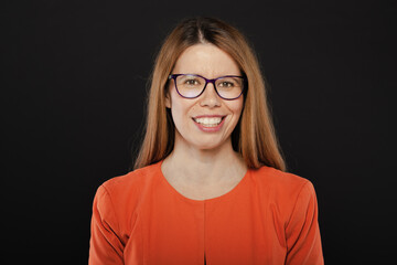 Portrait of a smiling Caucasian woman wearing an orange blouse and purple glasses, against a dark background, exuding confidence and happiness.