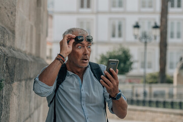 mature man with backpack looking at the phone with a surprised gesture on the street