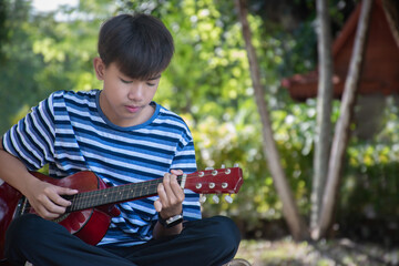 Asian preteen wearing headphones, sitting in local park, practising and playing ukulele and...