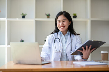 young asian lady doctor in white medical uniform with stethoscope using computer laptop talking...
