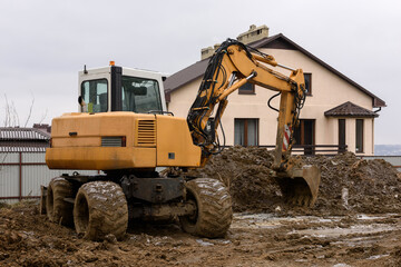 Obraz premium Yellow wheeled excavator at excavation work. A construction machine moves soil with a bucket or digs a pit for the foundation of a house. Construction site and construction of modern residential home.