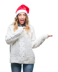 Beautiful young blonde woman wearing christmas hat over isolated background amazed and smiling to the camera while presenting with hand and pointing with finger.