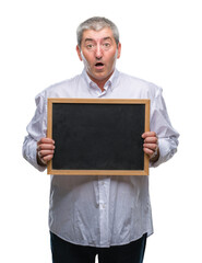 Handsome senior teacher man holding blackboard over isolated background scared in shock with a surprise face, afraid and excited with fear expression