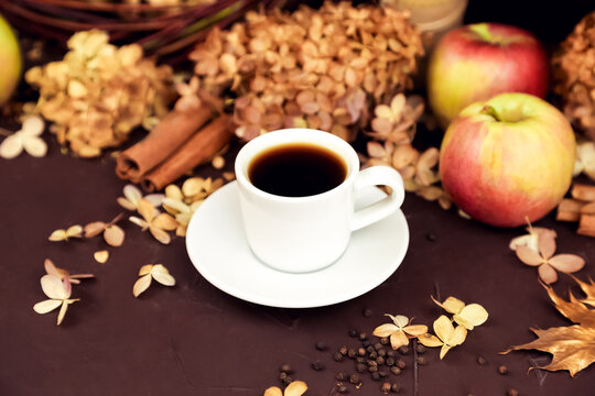 White coffee cup with espresso, cinnamon, red apples and dry hydrangea on dark brown background. autumn composition