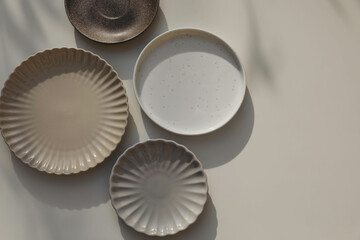 Different size and shapes of plates on beige table background in sunlight. Shadows. Neutral still life, web banner. Food, restaurant, craft concept. Scalloped pottery, ceramics. Flat lay, top view.