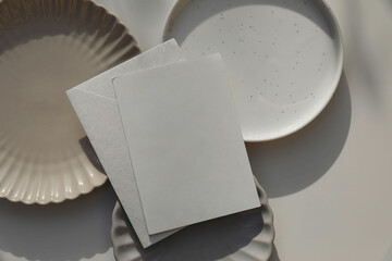 Blank greeting card, invitation mockup, envelope in sunlight. Different plates on beige table...