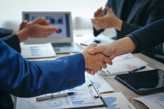 In a business team meeting, success is celebrated with a handshake. Hands close up, dressed in formal suits, working at desks with financial papers, calculators, and laptops.