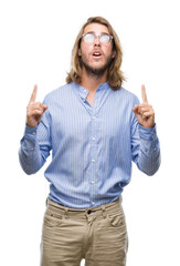Young handsome man with long hair wearing glasses over isolated background amazed and surprised looking up and pointing with fingers and raised arms.
