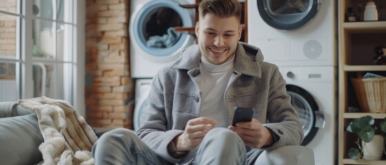 He loads dirty laundry in the machine while playing games on his smartphone. A handsome smiling young man sits in front of a washing machine while playing games on his smartphone.
