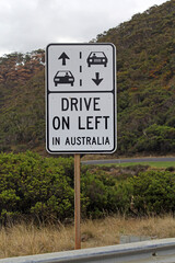 "Drive On Left In Australia" road sign amongst grass, shrubs and a mountain with trees