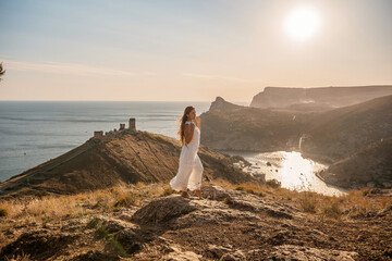 woman stands on a rocky hill overlooking a body of water. She is wearing a white dress and she is...