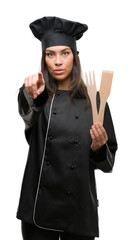 Young hispanic cook woman wearing chef uniform pointing with finger to the camera and to you, hand sign, positive and confident gesture from the front