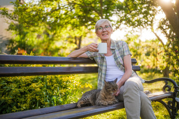 Happy senior woman enjoys reading book and spending time with her cat while sitting on a bench in...