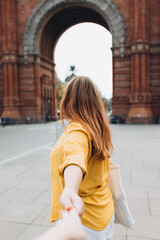 Woman with bag holding man by hand and going to Barcelona triumphal arch. Follow me, Young beautiful woman pulls the arm of her boyfriend in a city park