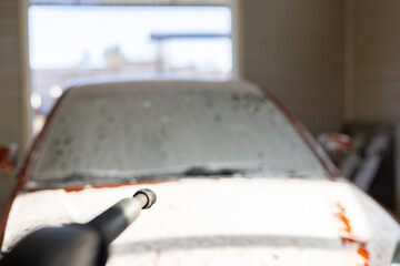 A man holds a gun in his hand at a car wash and covers his car with soap foam. The man took his car to a car wash to wash off all the dirt.