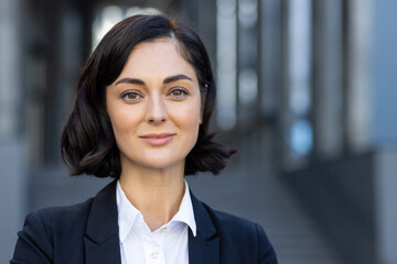 Fototapeta premium Close-up photo of a smiling young woman in a business suit standing outside a building and looking at the camera