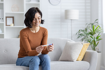 Happy young woman sitting on sofa at home and smilingly looking at home rapid pregnancy test, happy...