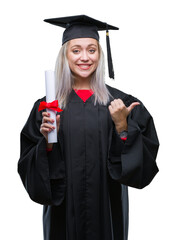 Young blonde woman wearing graduate uniform holding degree over isolated background pointing and...