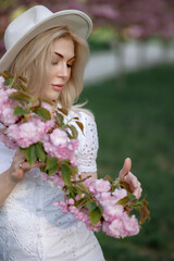 Obraz premium Beautiful young blonde woman in a hat holding a sakura branch in front of her