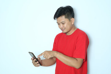 Fototapeta premium Young Asian man showing smiling while touching his mobile phone screen and looking at the mobile phone. Wearing red t-shirt