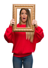Beautiful young woman holding vintage frame scared in shock with a surprise face, afraid and...