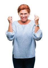 Atrractive senior caucasian redhead woman wearing winter sweater over isolated background excited for success with arms raised celebrating victory smiling. Winner concept.