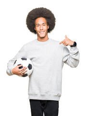 Young african american man with afro hair holding soccer football ball with surprise face pointing...