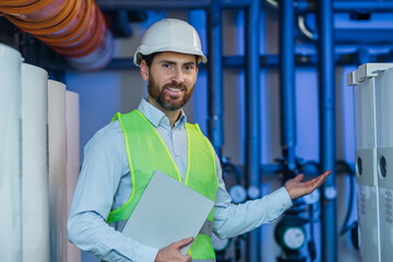 Portrait of male smiling worker in uniform and helmet with a laptop onn the background of boiler...