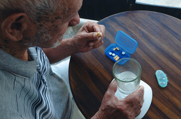 Hombre mayor nonagenario sentado sosteniendo una píldora y un vaso de agua con pastillero sobre...