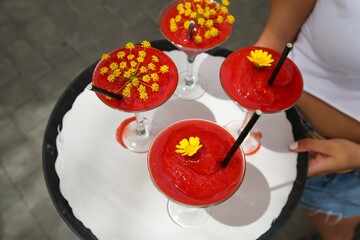A waitress holds a tray with beautiful red colored cocktails decorated with yellow flowers served with a black drinking straw