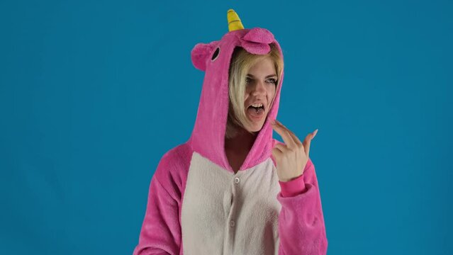 Female teenager in unicorn onesie makes vomiting gesture on blue background. Rebellious girl emotionally expresses disgust