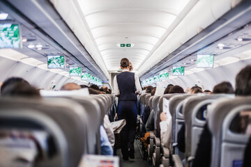 Interior of airplane with passengers on seats and stewardess in uniform walking the aisle, serving...