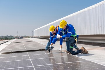 Two solar panel installation workers wearing safety gear are installing solar panels on a...