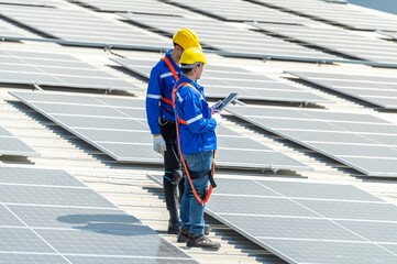 Two solar panel workers wearing safety gear are inspecting a large solar panel array on a commercial building. © BESTIMAGE