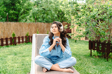 One young caucasian woman is relaxing in easy chair in her backyard while using her phone and listening to music on wireless headphones