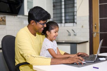 Obraz premium An Indian father smiles as he patiently helps his young daughter learn to use a laptop computer at their kitchen table