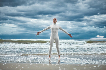 Happy hairless girl with alopecia in white futuristic suit stands with spread arms on beach bathed...
