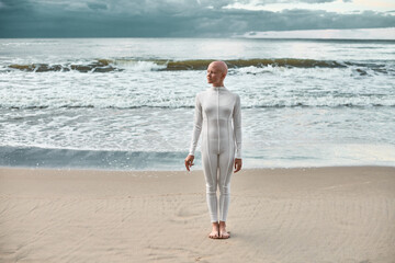 Full length portrait of young hairless girl with alopecia in white futuristic suit standing on sea...