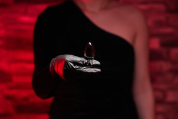 Metal anal plug in female hands in black latex gloves and a black dress on a red background. Sex...