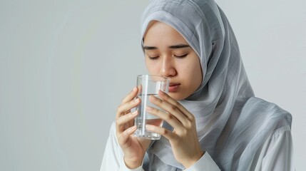 Muslim woman in hijab holding a glass of water and praying before drinking on a white background. Healthy lifestyle concept.