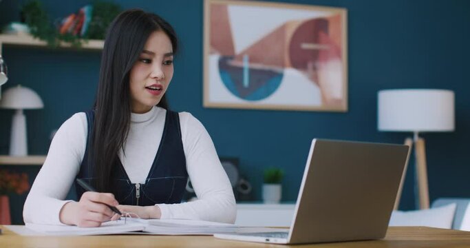 Hardworking young Chinese manager having videoconference with her boss or co-workers at home. Discussing work questions and looking directly into laptop camera while writing something in notebook.
