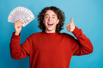 Photo of cheerful positive guy dressed red pullover rising money fan showing thumb up isolated blue...